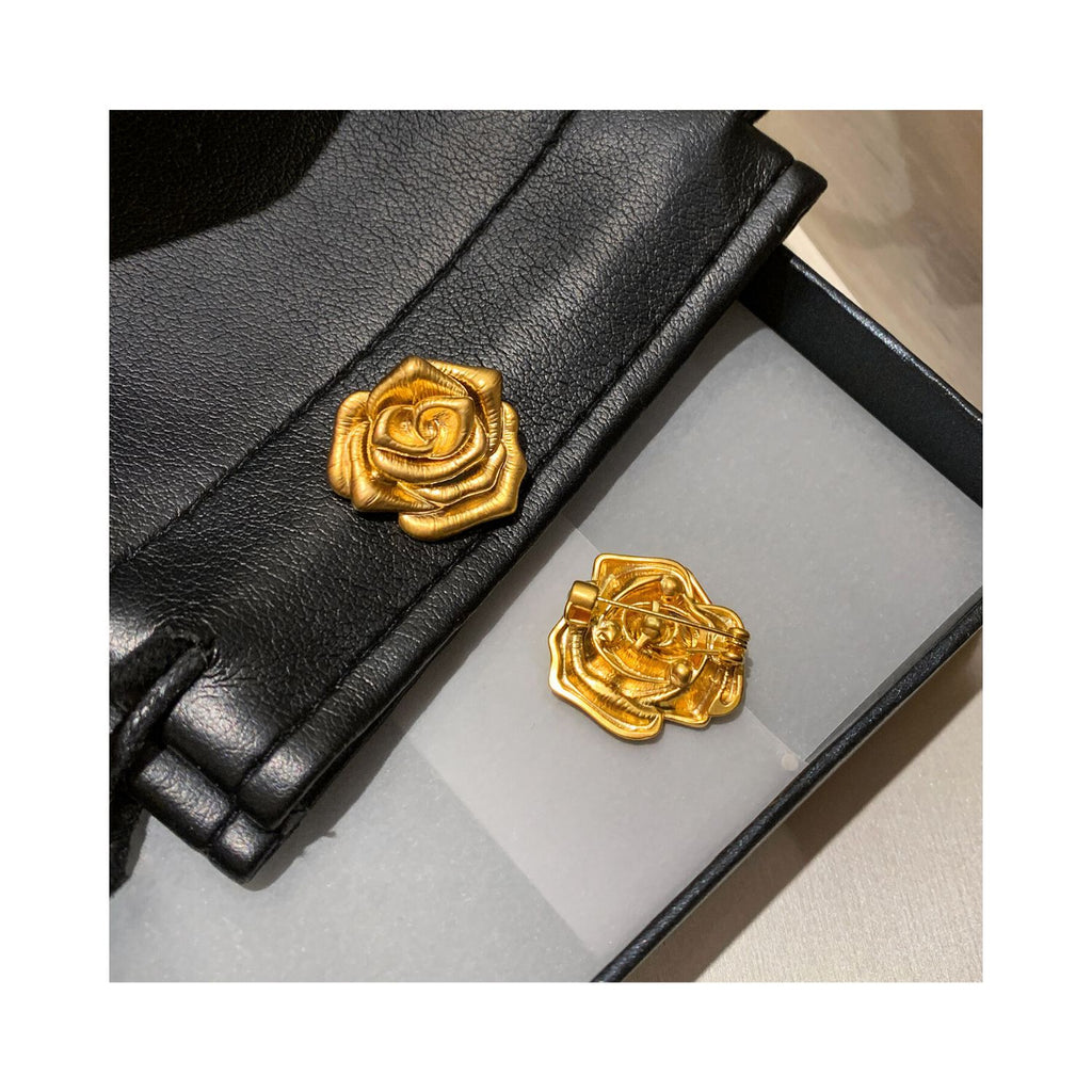 Flat lay of A Rose Is a Rose golden floral brooch with black leather accessories, highlighting the sculptural bloom form and versatile gift-ready styling.