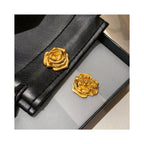 Flat lay of A Rose Is a Rose golden floral brooch with black leather accessories, highlighting the sculptural bloom form and versatile gift-ready styling.