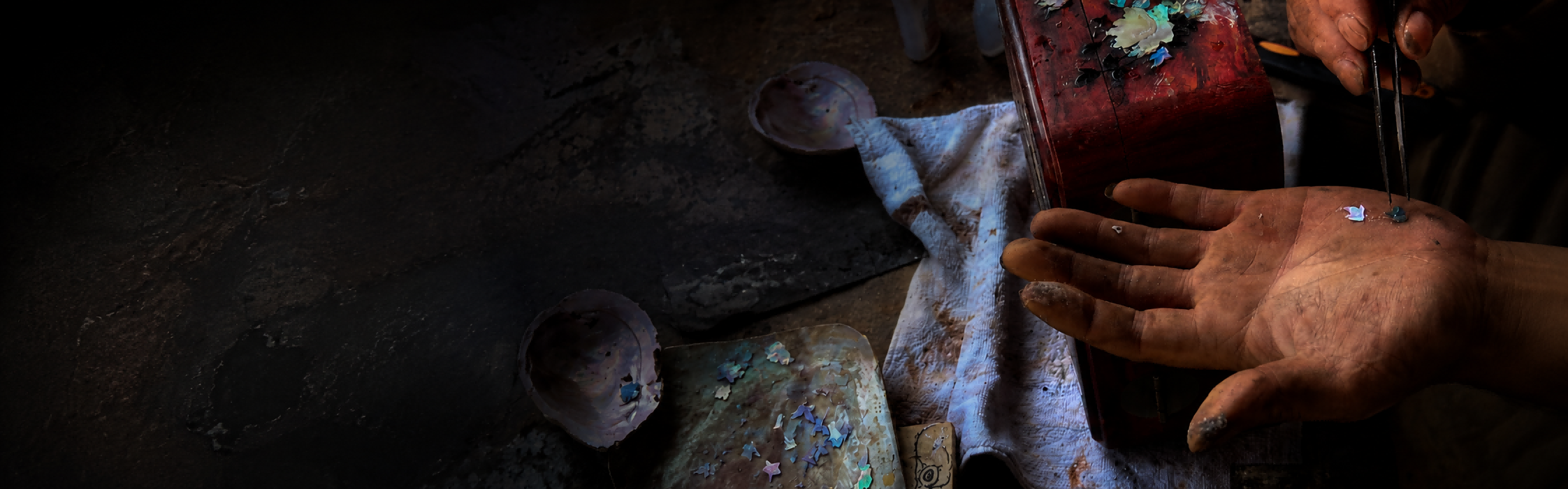 Jinherit About Us mission banner showing an artisan’s hands at a traditional workbench, surrounded by iridescent craft fragments, tools, and richly textured materials.