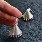 Jinherit Crimson Echo garnet bell drop earrings shown in hand, highlighting the tiered bell silhouette, spiral ridges, silver-tone texture, and deep red gemstone drops.