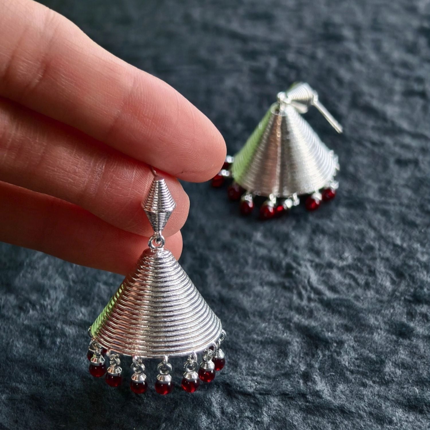 Jinherit Crimson Echo garnet bell drop earrings shown in hand, highlighting the tiered bell silhouette, spiral ridges, silver-tone texture, and deep red gemstone drops.