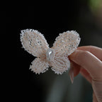 Jinherit Pearl Flutter hand-beaded butterfly hair clip shown in hand, featuring seed bead wings, a selected natural pearl centre, and a sculptural butterfly silhouette.