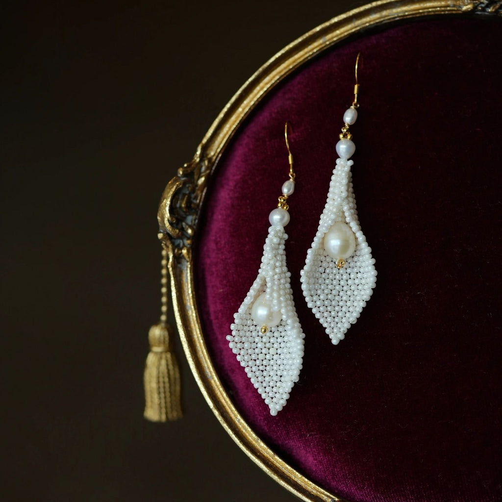 Jinherit Pure Calla white floral pearl drop earrings displayed on burgundy velvet, featuring hand-beaded calla lily forms, gold-tone findings, and softly luminous pearl details.
