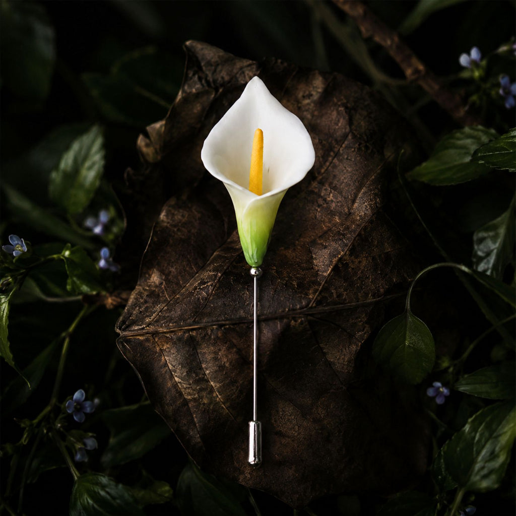 Jinherit Quiet Bloom ceramic calla lily brooch pin displayed on a dark botanical leaf background, featuring a white floral head, green detail, and slender lapel pin design.