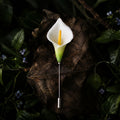 Jinherit Quiet Bloom ceramic calla lily brooch pin displayed on a dark botanical leaf background, featuring a white floral head, green detail, and slender lapel pin design.