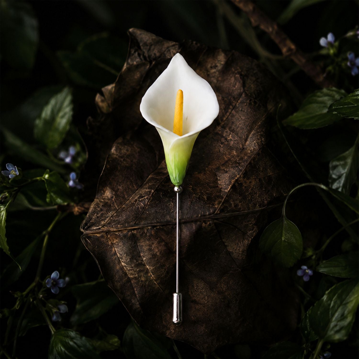 Jinherit Quiet Bloom ceramic calla lily brooch pin displayed on a dark botanical leaf background, featuring a white floral head, green detail, and slender lapel pin design.