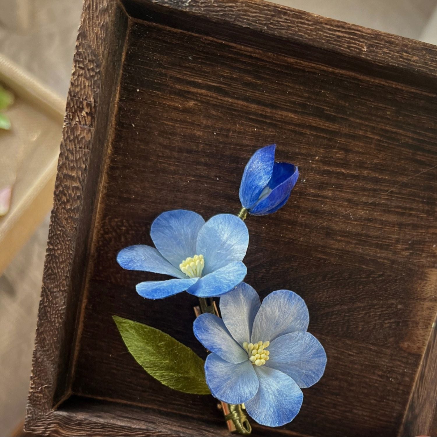 Jinherit Sky Bloom Ronghua silk flower hair clip displayed in a wooden tray, featuring hand-shaped blue mulberry silk blossoms, green leaf accents, and a flat alligator clip.