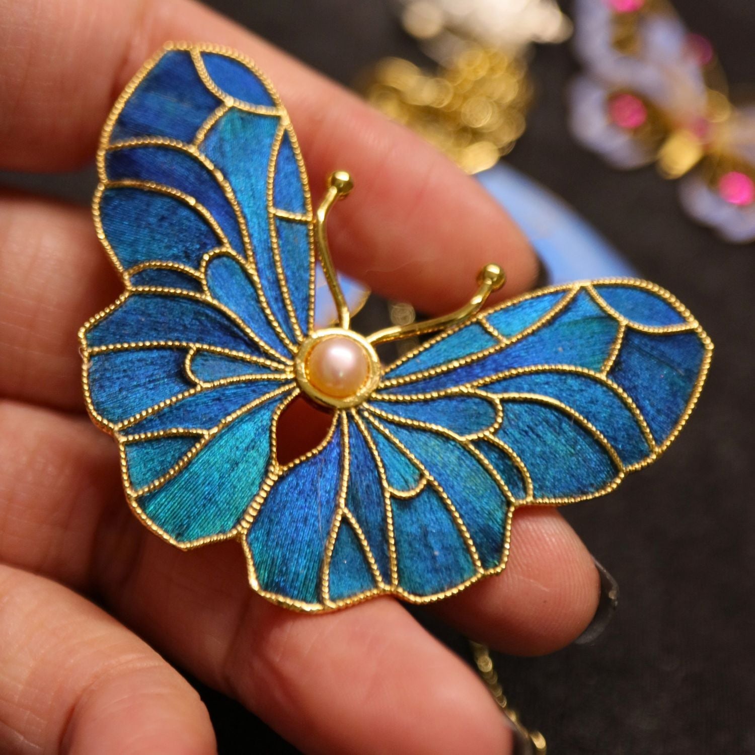 Hand holding Dian Cui-inspired butterfly brooch showing scale, feather texture, and pearl centre detail.
