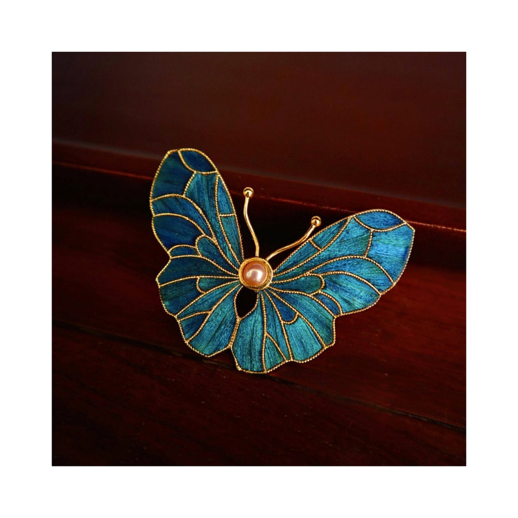 Angled front view of Whispering Wings Dian Cui butterfly brooch on a wooden surface, showing vivid blue iridescence and layered wing shape.