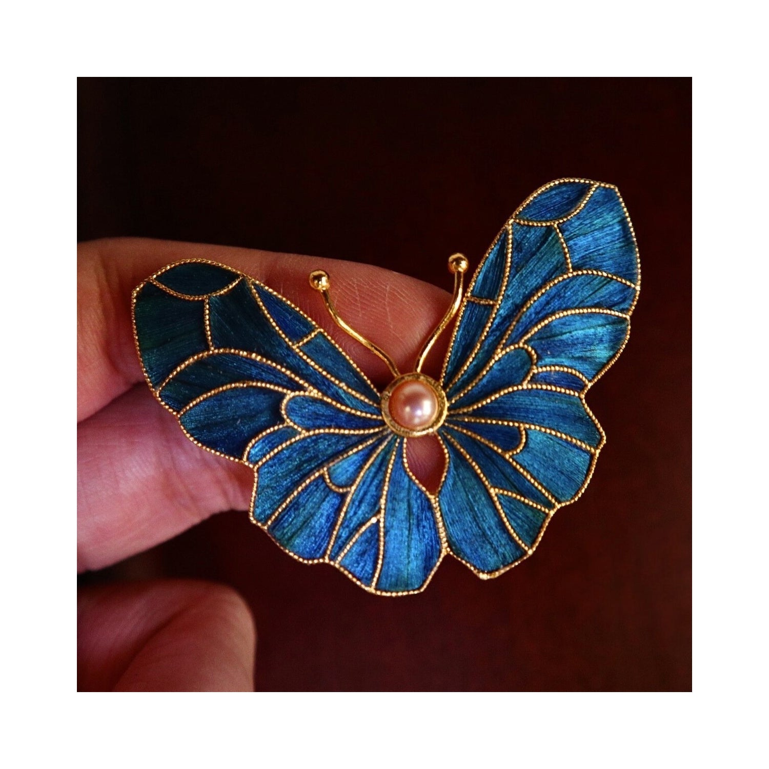 Handheld view of Whispering Wings Dian Cui butterfly brooch showing artisanal scale, feather texture, and luminous blue wing panels.